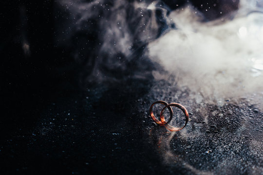 Wedding rings on dark surface shining with light close up macro. Water splashes. Reflection. Lights and smoke.