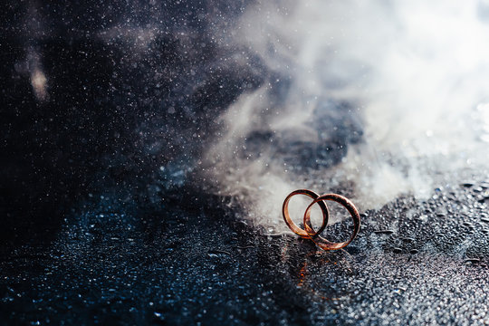 Wedding rings on dark surface shining with light close up macro. Water splashes. Reflection. Lights and smoke.