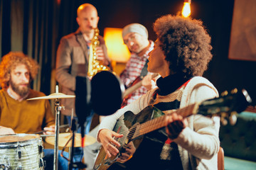 Mixed race woman singing and playing guitar while sitting on chair.In background drummer, saxophonist and bass guitarist.