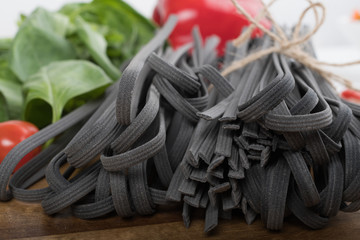 Black squid ink tinted tagliatelle pasta, uncooked, with basil and tomatoes in the background, on a...
