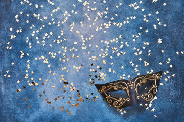 Black carnival mask and golden stars on blue background. Top view, copy space.