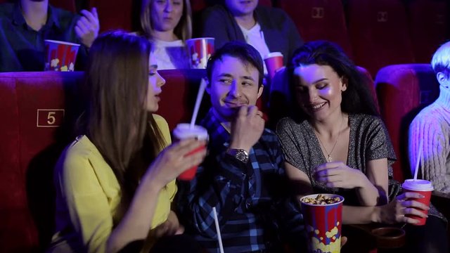 A young guy sits in a cinema with two girls and flirts with them. A girl gives a drink to her cola young guy in the cinema. Young man on a date in a cinema with two girls.
