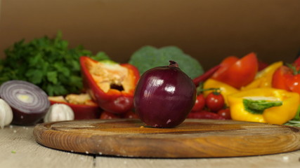 red onions in half on a wooden board, the bow cut into two halves is swinging on a wooden board, in the background vegetables, greens, garlic, peppers, tomatoes, brocols, side view