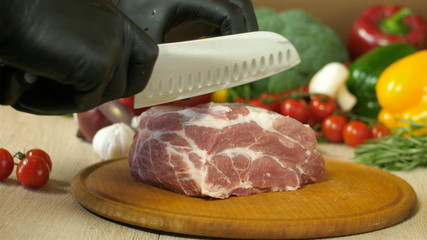 Chef with black gloves takes a knife and regulates a piece of raw juicy meat lying on a round wooden board, next there is another round wooden board to a piece of meat, in the background vegetables