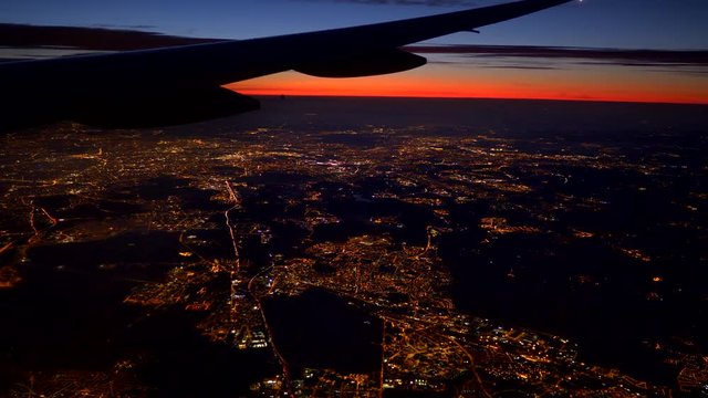 Flying over lights of big city. Aerial sunrise view from plane porthole. 4K UHD