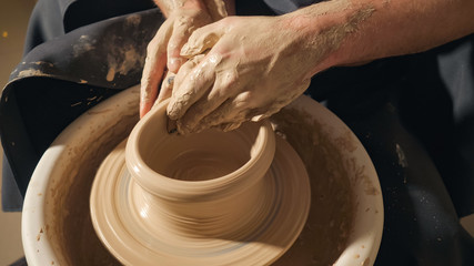 Two intertwined hands form a pot of clay on a potter's wheel. A male hand helps a woman's hands shape a clay shape.