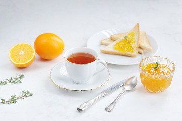 Toasts, orange jam and a cup of tea. The concept of table setting for breakfast. The composition is complemented by oranges and cutlery. Light background. Close-up.