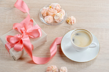 The gift is tied with a ribbon of coral color, a cup of coffee and meringue on a light wooden surface. The concept of receiving a gift on a holiday. 