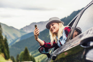 Smiling young woman taking selfie picture with smart phone camera outdoors in car