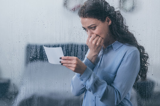 grieving woman holding photograph, covering mouth with hand and crying at home through window with raindrops