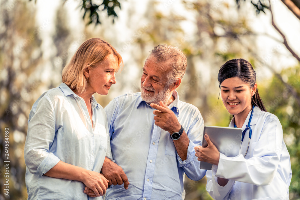 Wall mural Senior couple man and woman talking to young nurse or caregiver in the park. Mature people healthcare and medical staff service concept.