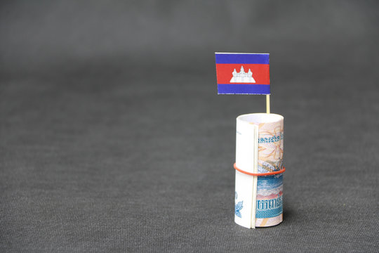Rolled banknote money one thousand Cambodian Riel and stick with mini Cambodia flag on dark grey floor and background.