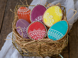 Colored cookies in the form of Easter beautiful eggs with icing lace in a metal golden basket with straw on wooden table. Close up, selective focus, copy space.