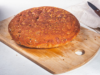 Homemade round bread made of coarse flour sprinkled with white and black seeds of sesame seeds lies on a kitchen wooden board and a white table.