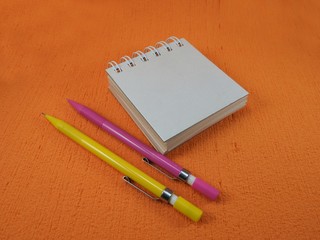 A small blank notepad with two mechanical pencils (one yellow and one pink) on an orange surface. The notepad is closed.
