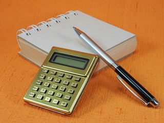 Close-up of a golden calculator, a small blank notepad and a pen on an orange surface. The notepad is closed.