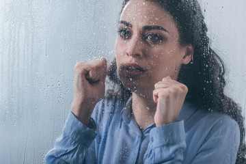 beautiful sad woman with clenched fists looking through window with raindrops