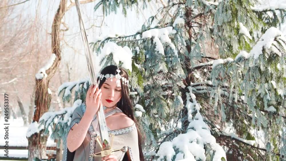 Wall mural woman fantasy warrior holding royal elven sword in hand. long black hair, red lips make-up posing for camera, elf princess with blade sharp weapon. Creative dress gray cloak in winter forest pine tree