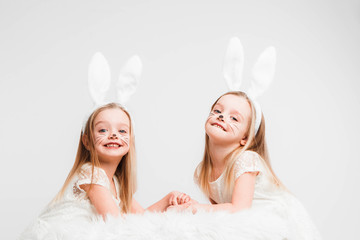 Little blonde twins in white dresses with rabbit ears. Studio photo on gray background. Kids celebrate Easter.