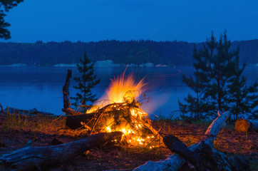 Big night bonfire at the shore of the river in a forest glade, flames, sparks.