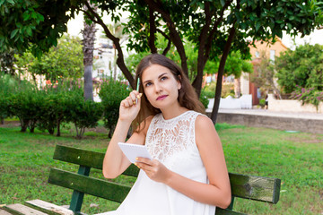 A beautiful woman who is holding a notebook and a pen up has some idea and is thinking about something on the bench in the park