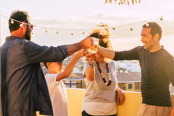 Group of friends toasting and clinking glasses with beer and orange juice together during the sunset on the roof terrace with city view - mixed ages and generations having fun celebrating