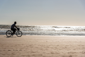 Fototapeta premium Silhouette of man riding bicycle on a beach with the sun reflecting in the sea and a clear sky