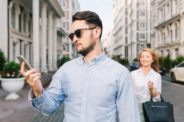 Portrait of a little bit sad gut in sunglasses looking on phone on street. Pretty blonde girl catching him from behind on background