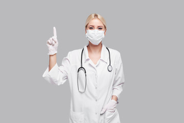 Beautiful young doctor woman in medical white coat and a protective mask putting on rubber gloves. Isolated over grey background. Copy space for text