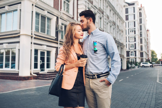 Lovely Couple Walking Around British Quarter. Dark-haired Man In Blue Shirt Kissing In Head Blonde Girl In Black Dress With Coral Jacket