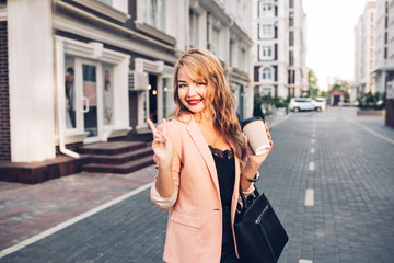 Portrait fashionable blonde woman with long hair walking in coral jacket on street. She holds a cup of coffee, smiling to camera