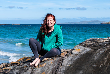 Woman wearing a turquoise hoodie with red hair sitting on some rocks with beach and ocean in the background. Taken on Renvyle beach, along the Wild Atlantic Way in Ireland in summer.