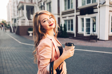 Fototapeta premium Closeup portrait attractive model with vinous lips walking with coffee in coral jacket on street. She smiling to camera