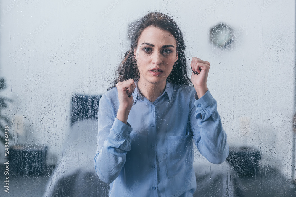 Wall mural beautiful sad woman with clenched fists looking at camera through window with raindrops - Wall murals
