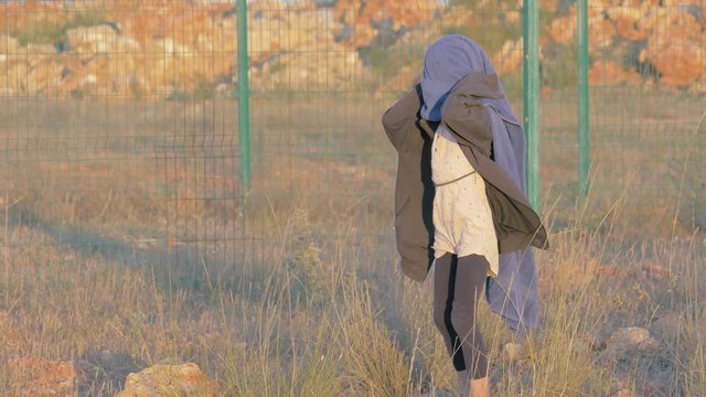 Unhappy strange little girl walking refugee camp covered from cold with blue blanket. border area place of accommodation migrants child playing background fence with barbed wire. concept lifestyle