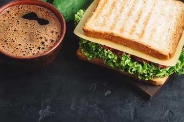 Rolgordijnen Cup with hot coffee and sandwich with grilled toast, salami sausage, salad lettuce, spinach leaves and cheese on a dark background © O.Farion