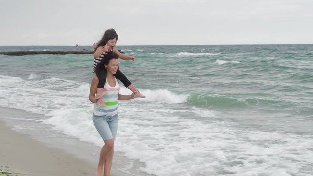 Walk with the child on the beach. Fun with mom on the beach. Happy mother with her daughter on her shoulders are having fun on the background of the sea.