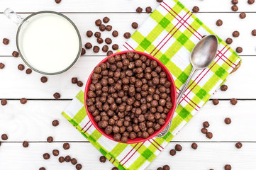 Bowl with chocolate corn balls, spoon and cup of milk on white wooden table. Top view.