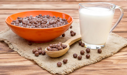 Bowl with chocolate cereal balls in milk, cup of milk and wooden spoon on sack napkin.