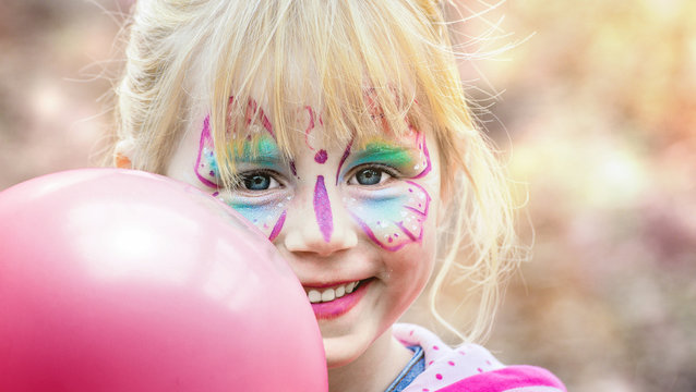 Glückliches geschminktes Mädchen beim Kinderkarneval auf einem Freizeitpark mit einem pinken Luftballon