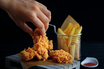 Hand grabs crispy kentucky fried chicken. French fries and tomato on black wooden table