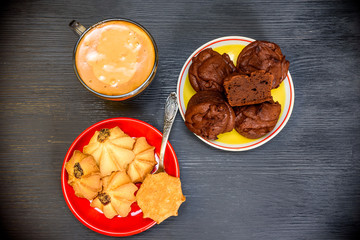 Chocolate muffins on a plate, shortbread and a cup of coffee photographed close-up on a black wooden background.