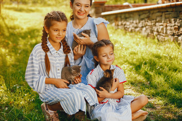 Fototapeta premium three beautiful and cute girls in blue dresses with beautiful hairstyles and make-up sitting in a sunny green garden and playing with a cats