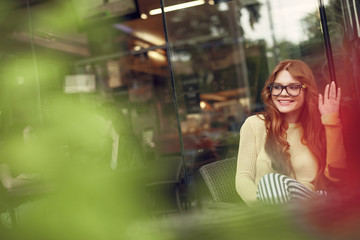 Attractive red-haired girl in glasses showing hello gesture in cafe