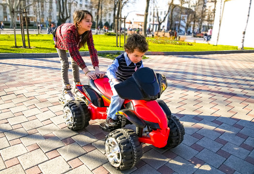 Kids driving electric toy car in urban summer park. Outdoor toys. Children in battery power vehicle. Little boy riding toy truck. Girl riding on roller skates cling to the car