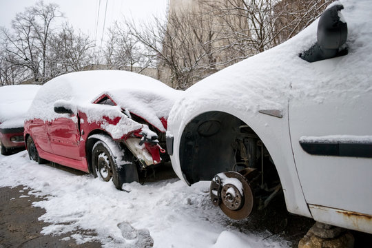 Two damaged cars, one red and one white, covered with snow, sitting in front of each other in a line of broken cars, awaiting repair inside an auto service courtyard - Bucharest, Romania