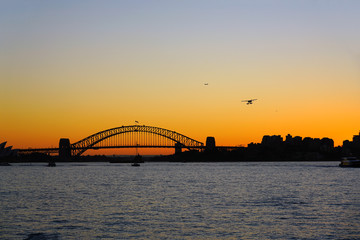 Fototapeta na wymiar Sunset view of a seaplane flying in the orange sky by the iconic steel Sydney Harbour Bridge in New South Wales, Australia