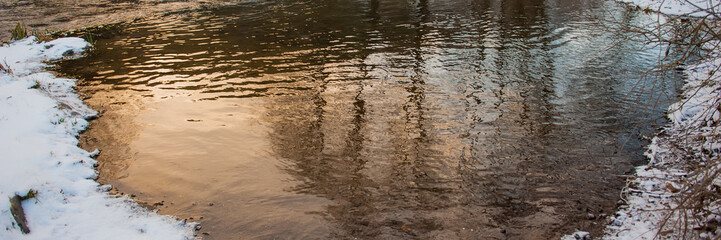 river bank covered with snow and reflection with waves on the surface of the water in the river.