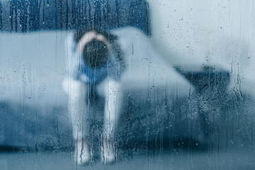 depressed woman sitting on bed and holding head in hands through window with raindrops - obrazy, fototapety, plakaty