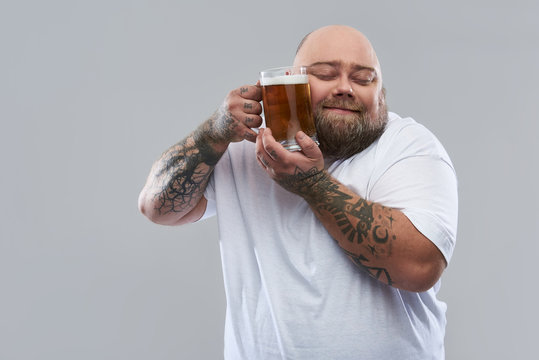 Waist up of bearded tattooed man pressing glass of beer to his cheek